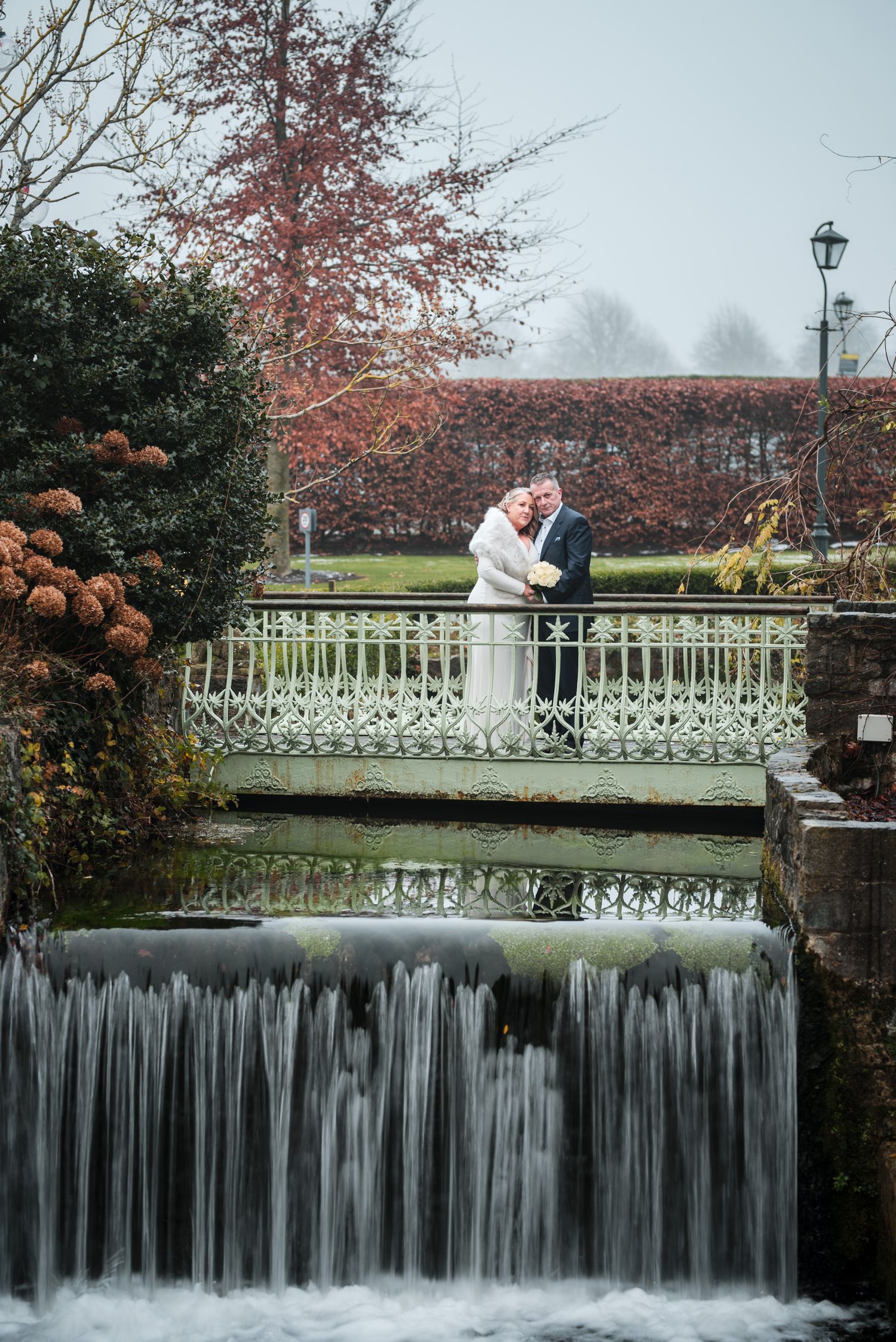 Couple embraces in romantic wedding moment against scenic waterfall backdrop with autumn foliage and white railing bridge.