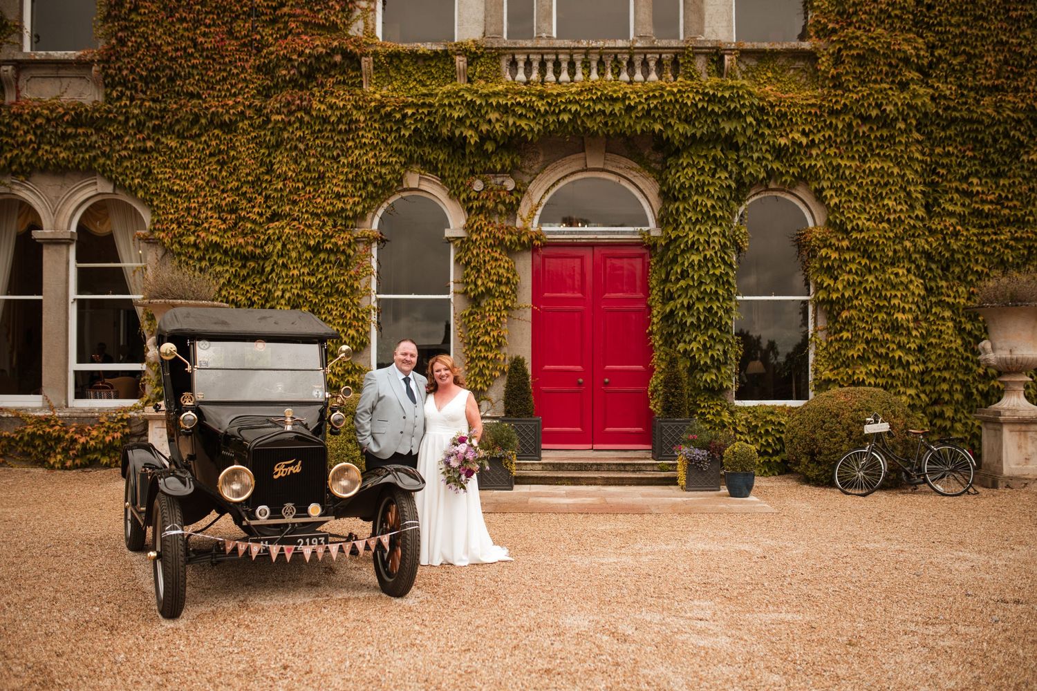 Vintage Model T Ford parked in front of ivy-covered mansion with red door and couple in wedding attire on gravel drive.