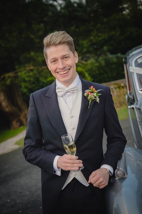 A groom in a formal navy tuxedo with bow tie and boutonniere holds a champagne glass beside a classic car.