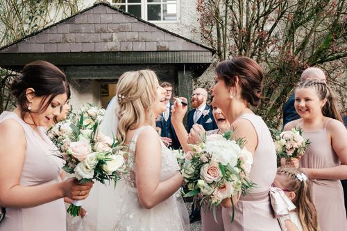 Wedding party in blush pink dresses share joyful moment while holding white floral bouquets outside house.