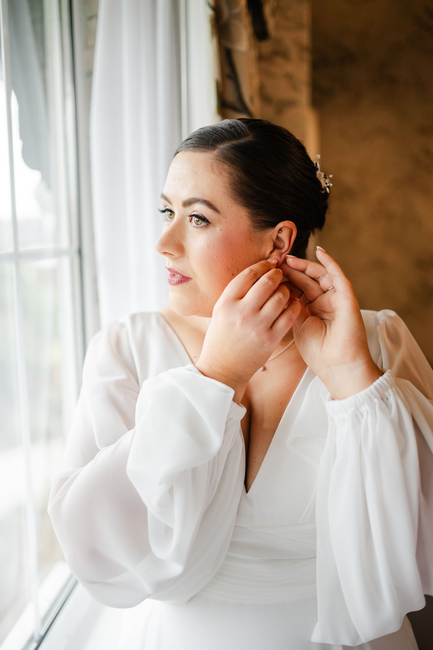 Someone in a white bridal gown with billowing sleeves puts on earrings by a window.