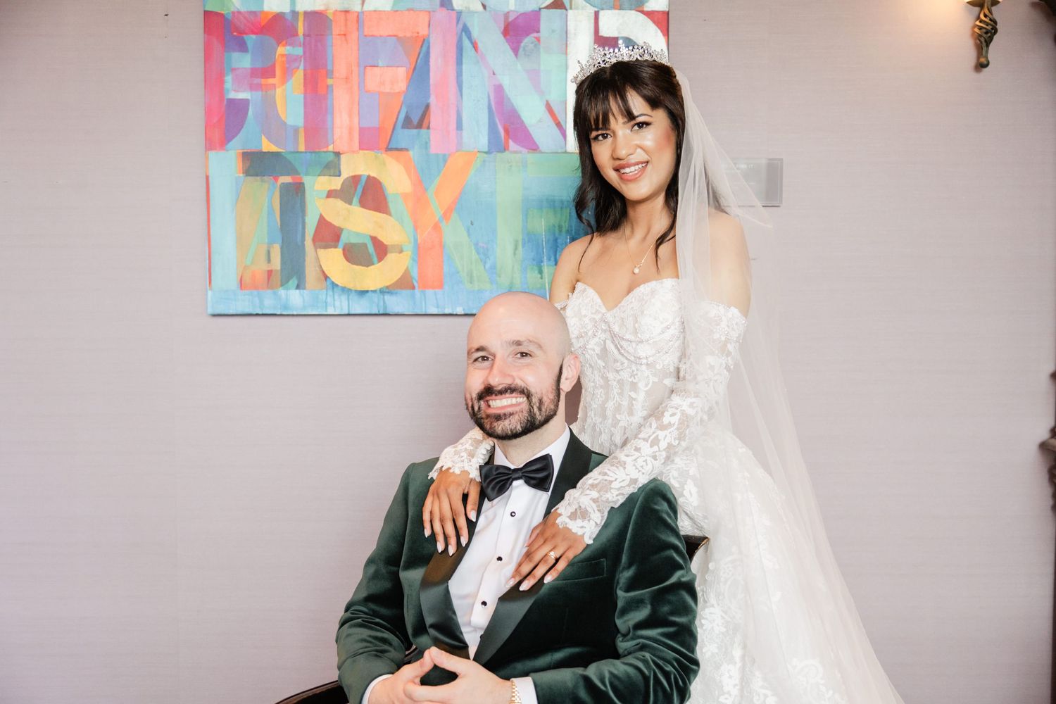 Bride in lace gown and groom in green velvet tuxedo pose together at wedding reception with colorful art backdrop.