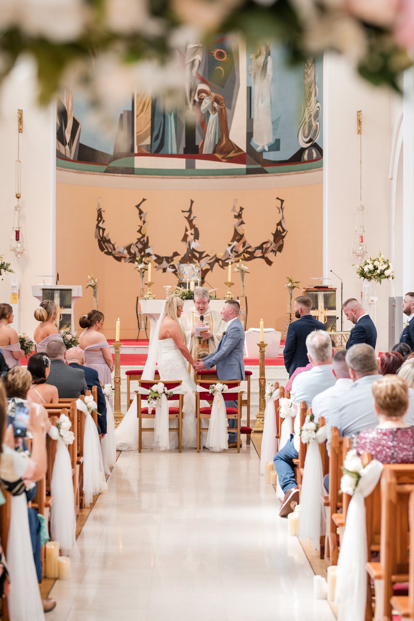 A church wedding ceremony in progress with guests seated in wooden pews decorated with white bows and floral arrangements.