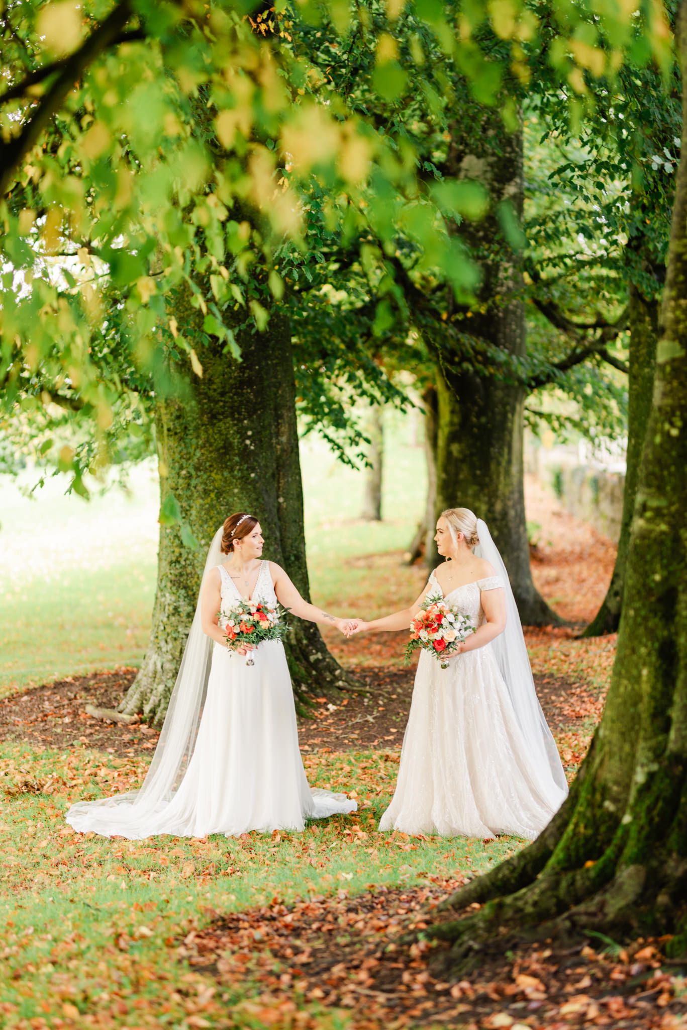 Two figures in flowing white wedding gowns and veils hold bouquets while standing among tall trees on autumn ground.