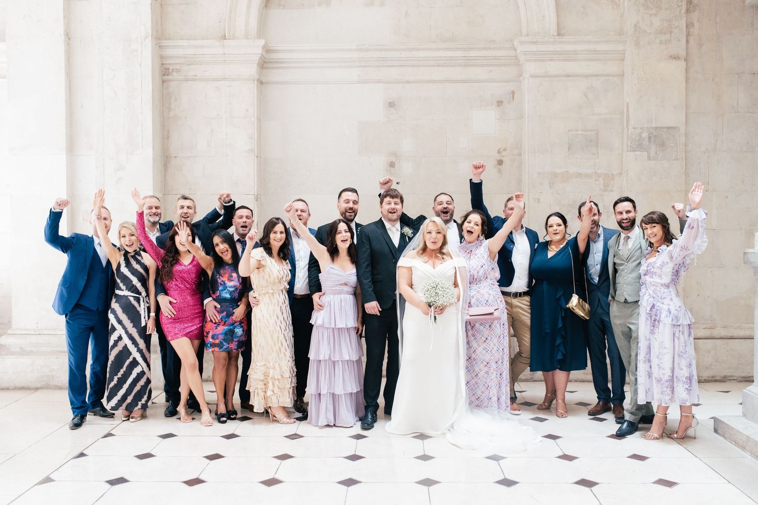 A group of wedding guests and party members celebrating with raised arms in front of a light stone building.