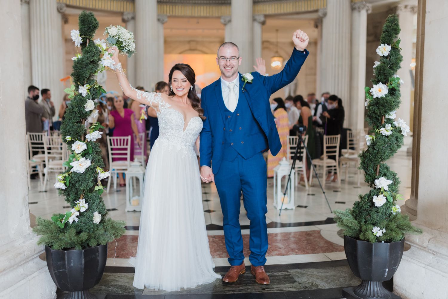 A couple celebrates at their wedding ceremony under an elegant archway with floral arrangements and white roses.