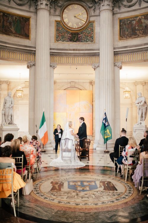 Wedding ceremony taking place in an ornate circular hall with marble columns, classic statues and flags of Ireland.