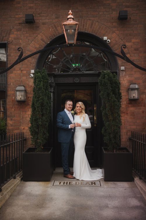 Elegant couple stands in brick archway entrance with lanterns and greenery at The Dean venue.