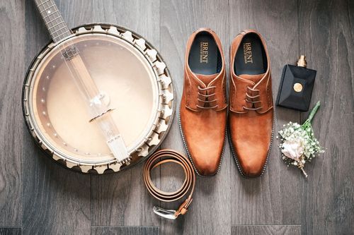 A flat lay of groom's wedding accessories including brown leather dress shoes, belt, watch, and boutonniere on dark wood floor.