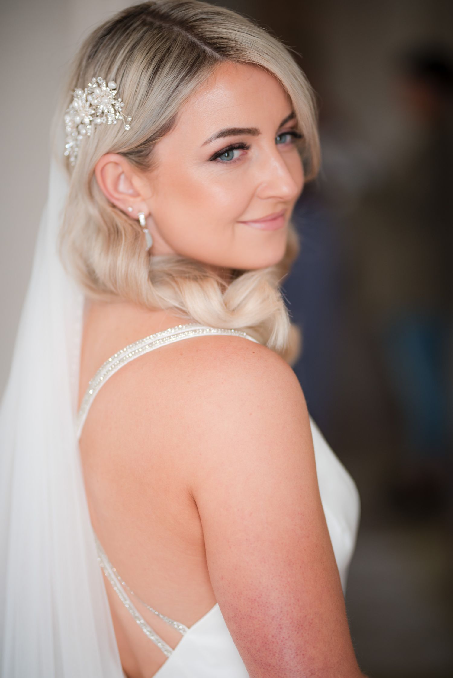 Elegant bride in white dress with flowing veil and delicate hair accessory gazes over shoulder with a gentle smile.