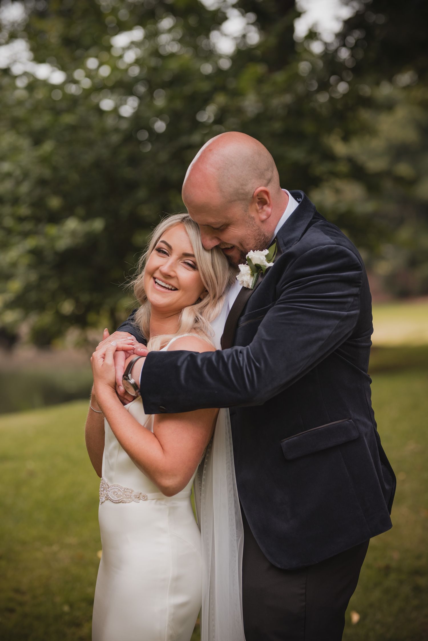 Candid moment between bride and groom embracing outdoors with natural bokeh background.