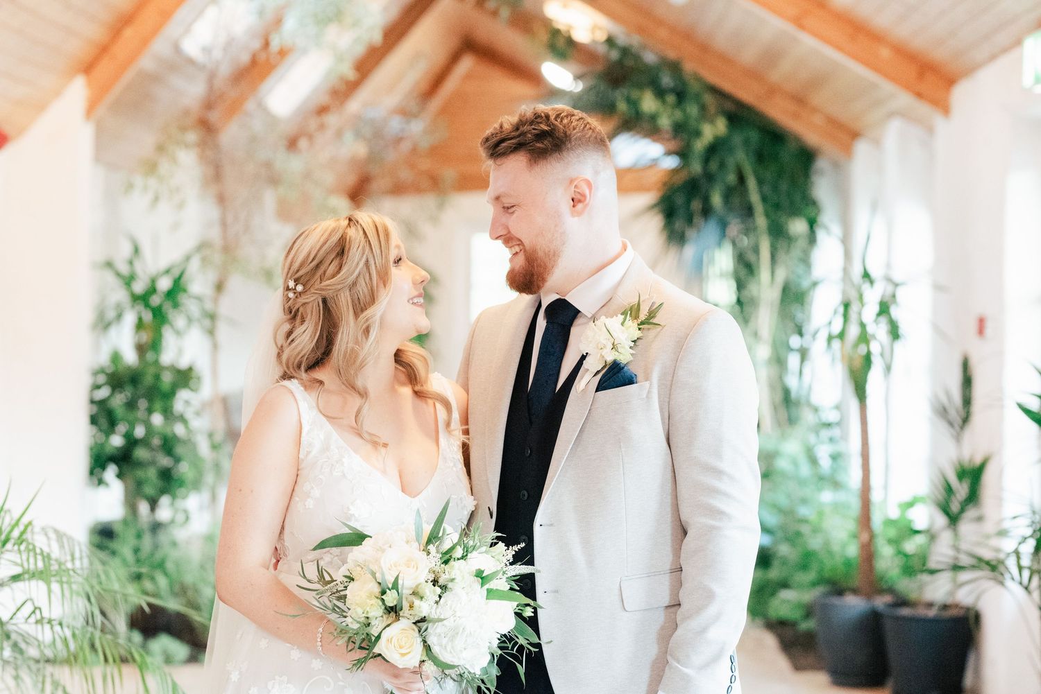 Bride and groom share loving glance in bright indoor venue with tropical plants and wooden beams.