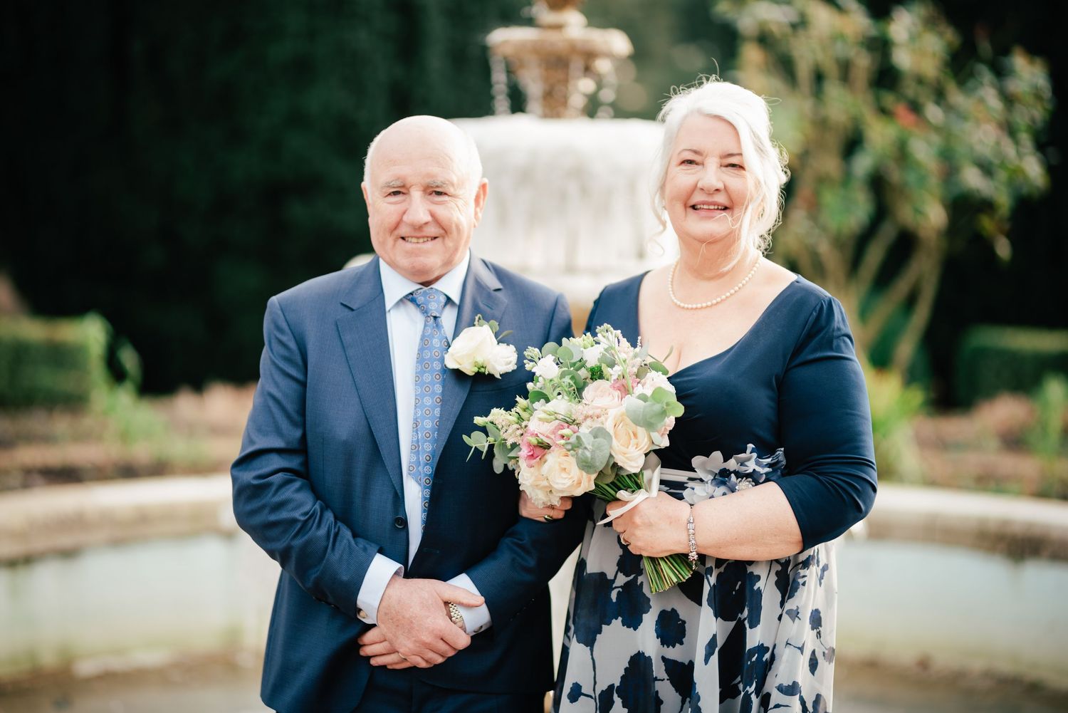 A joyful couple stands together outdoors near a fountain in formal attire during a wedding celebration.
