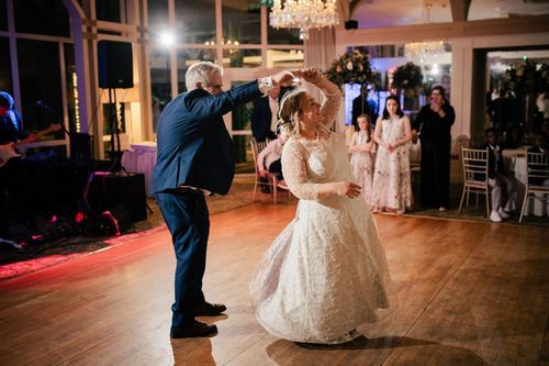Two people dance together at a wedding reception on a wooden dance floor under an elegant chandelier.