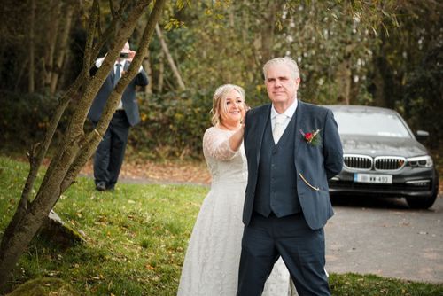 Couple celebrates their wedding day outdoors in autumn, with a photographer visible in background near parked car.
