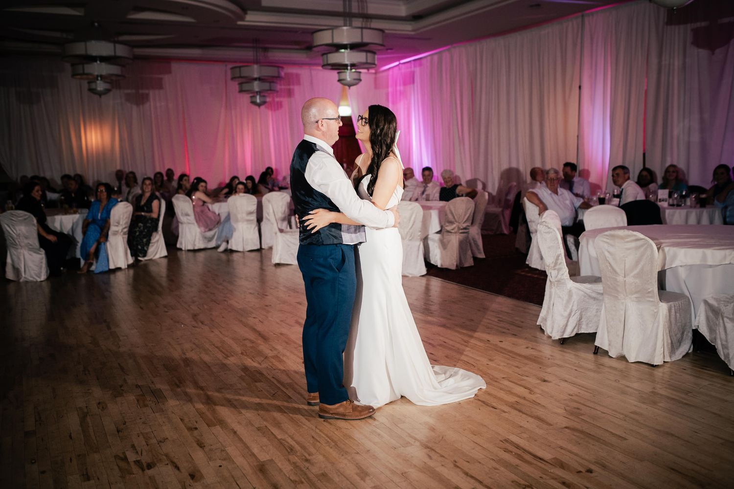 Couples dancing on wooden floor at wedding reception with pink uplighting and white tablecloths in background.