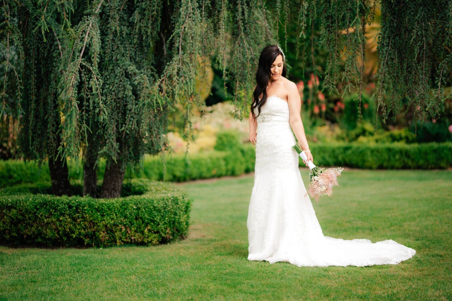 A bride in a flowing white gown with train stands in a garden with weeping willows and manicured hedges.