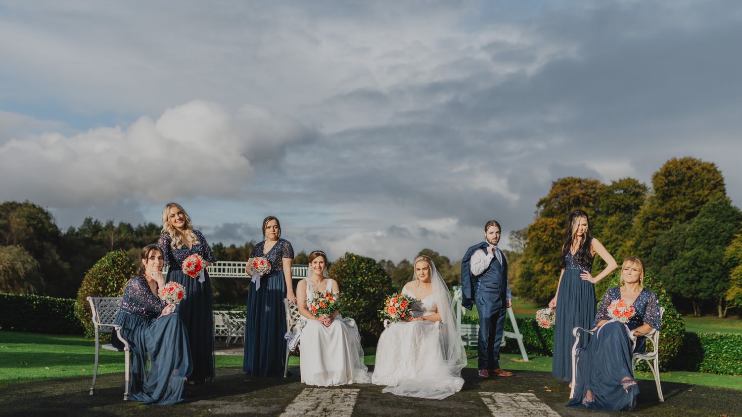 Wedding party in blue dresses and suits posing outdoors for photos in a sunny garden setting.