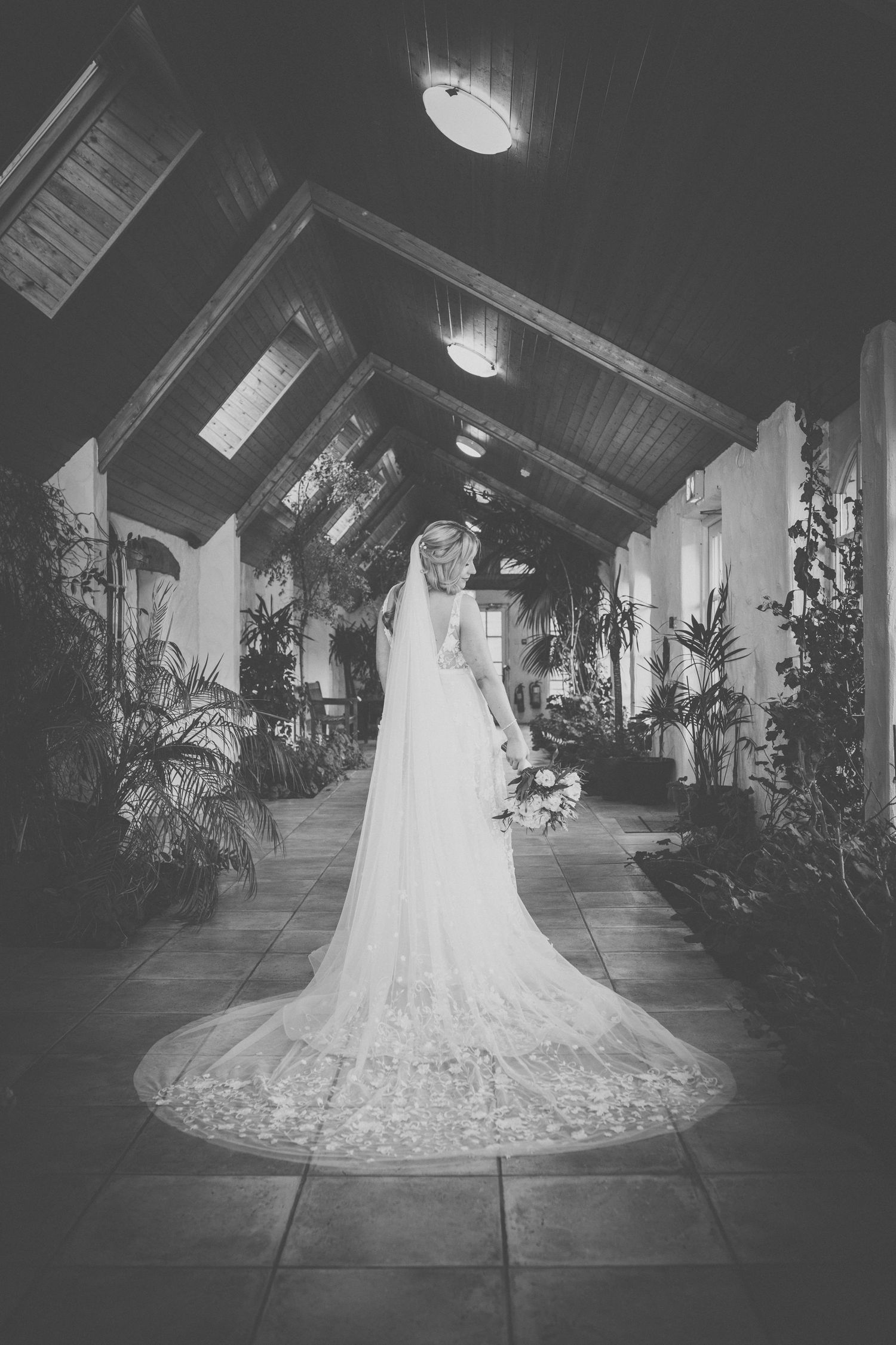 Black and white photo of bride in lace wedding gown standing in dramatic hallway with trailing veil.