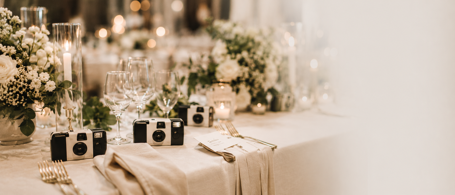 Elegant wedding reception table with disposable cameras, white floral centerpieces, candles, and neutral linen runner.