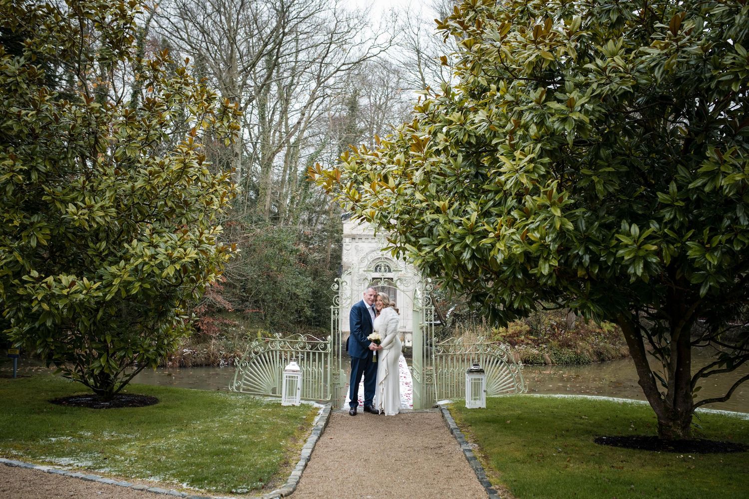 A couple walks down a garden path lined with lanterns and trees on an overcast day.