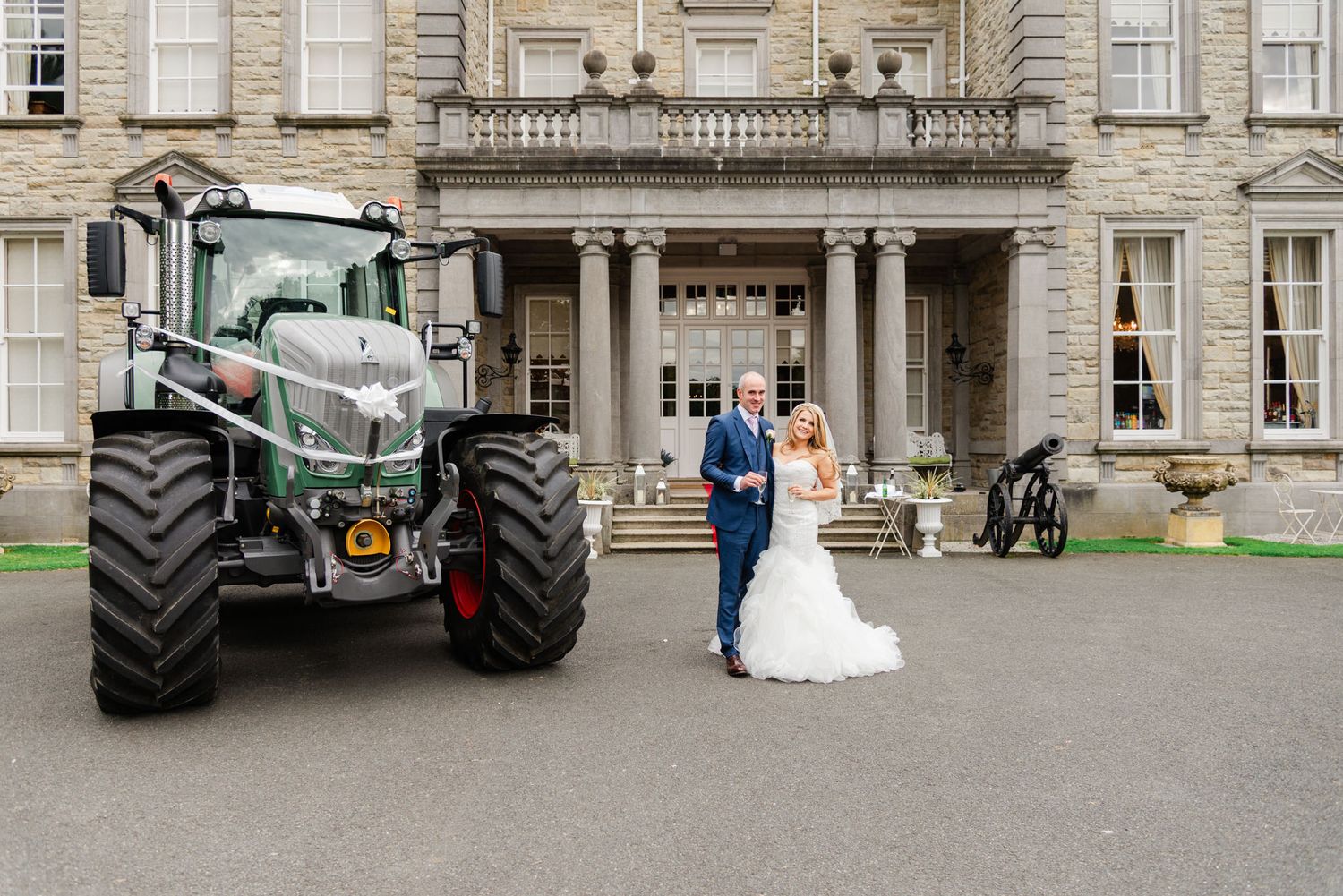 A couple in wedding attire stands between a green farm tractor and an elegant stone mansion for a wedding photo.