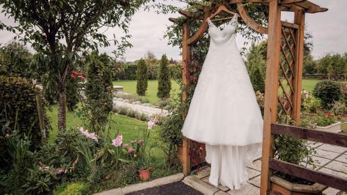 A white lace wedding dress hangs from a wooden garden arbor overlooking a scenic landscape with trees and paths.