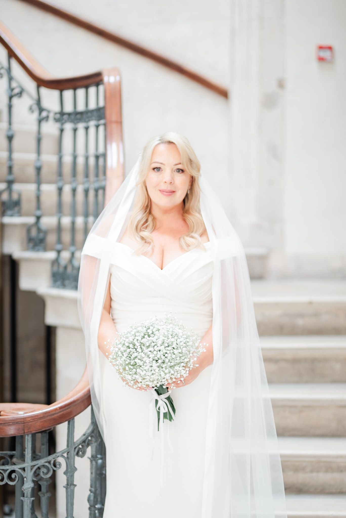 A bride holding a baby's breath bouquet poses on a classic staircase in her wedding gown.