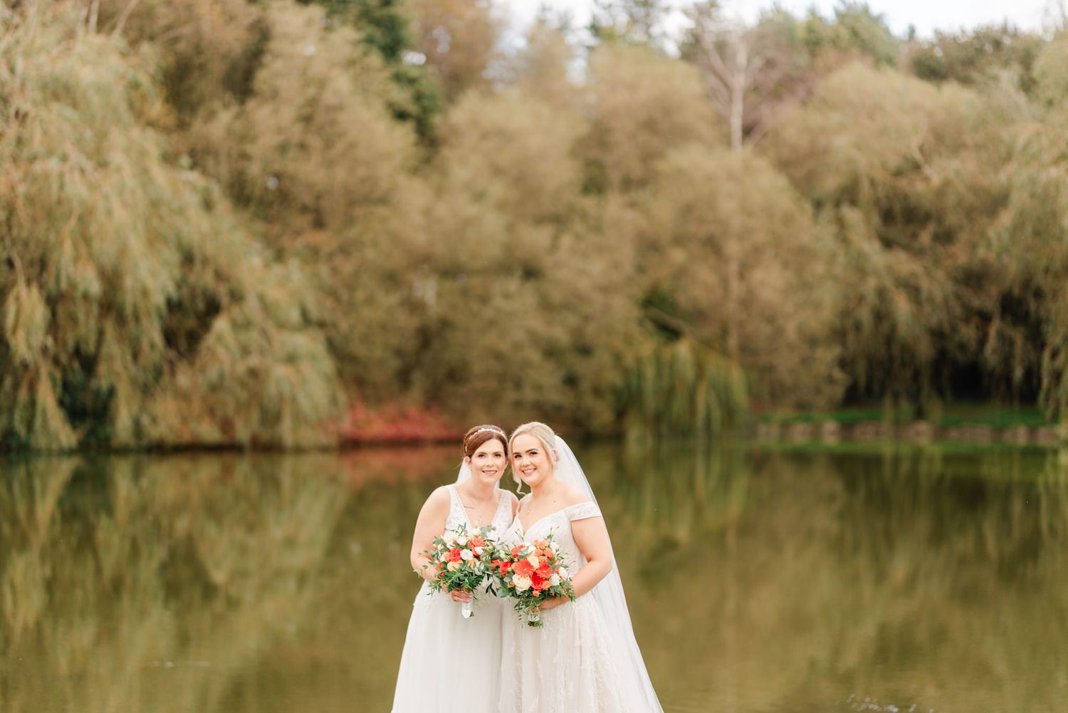 Two brides in white dresses stand together by a serene lake surrounded by autumn trees.
