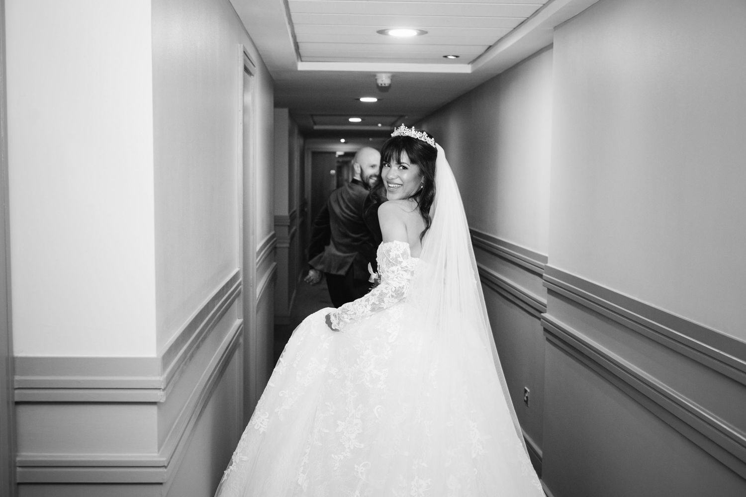 Black and white photo of a bride in a lace gown sharing a tender moment with a guest in a hotel hallway.