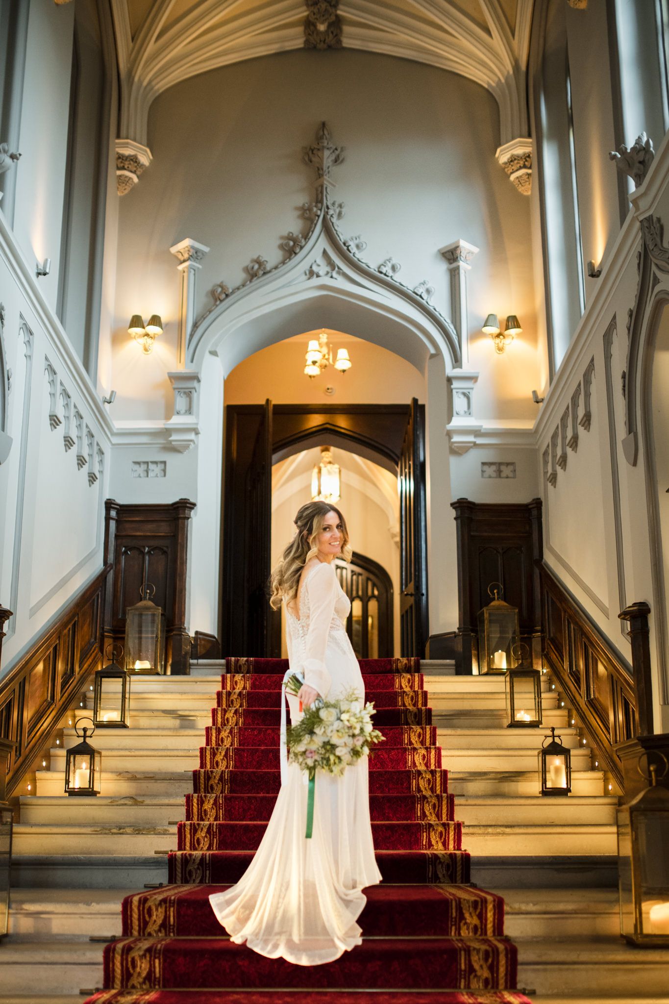 Elegant long-sleeve wedding gown on grand staircase with red carpet and Gothic arches illuminated by lanterns.