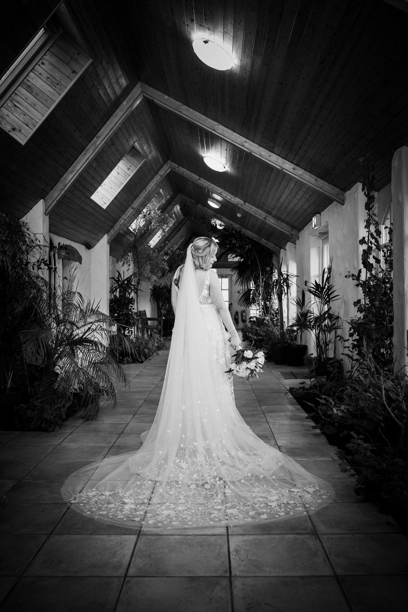 Elegant white wedding dress with intricate lace details displayed in dramatic black and white along a hallway.