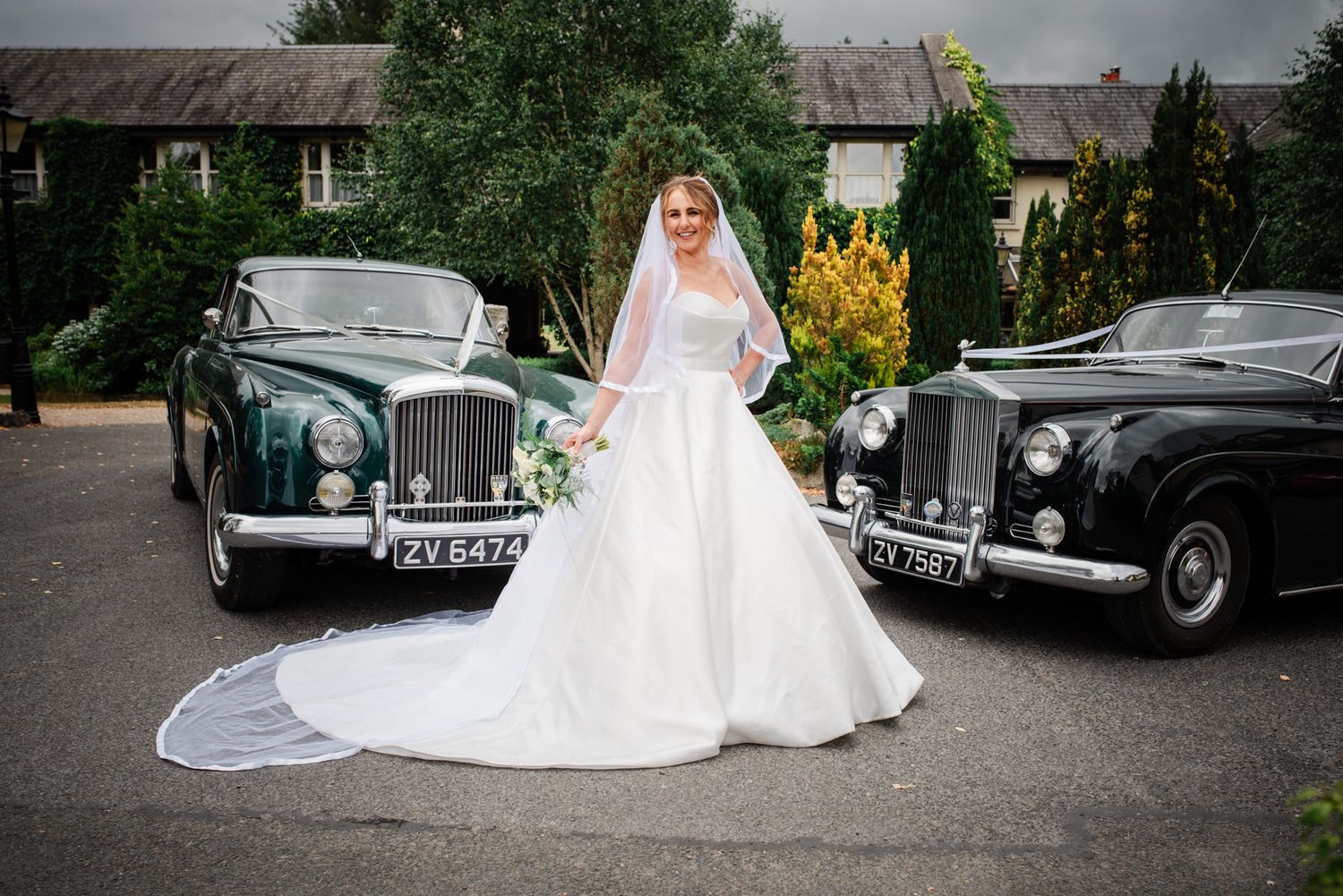 White bridal gown with long train positioned between two classic vintage black luxury cars.