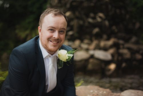 A person in a dark suit and white tie smiles outdoors while wearing a white rose boutonniere.