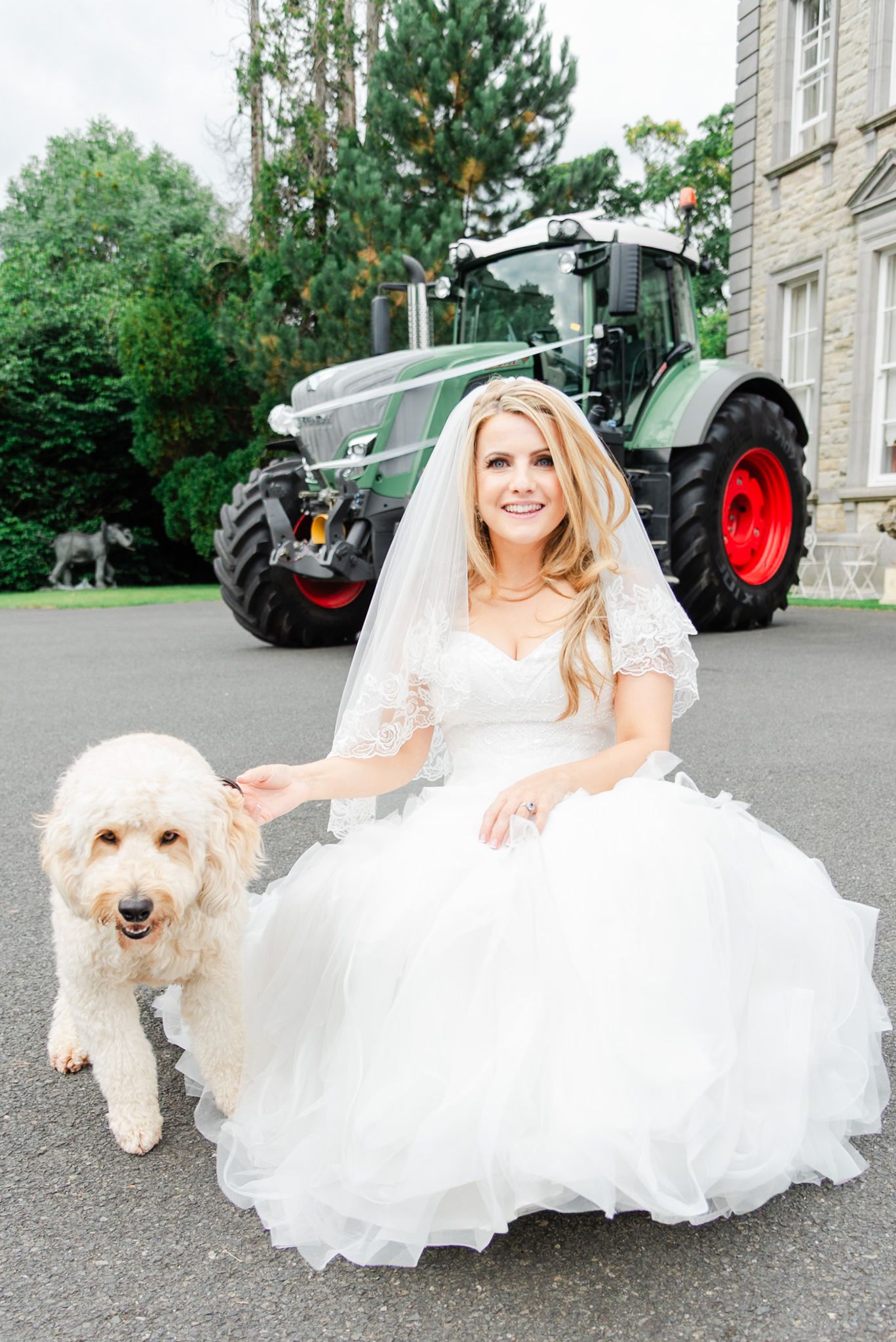 A bride in a white wedding dress poses with a fluffy white dog next to a large green tractor on a country estate.