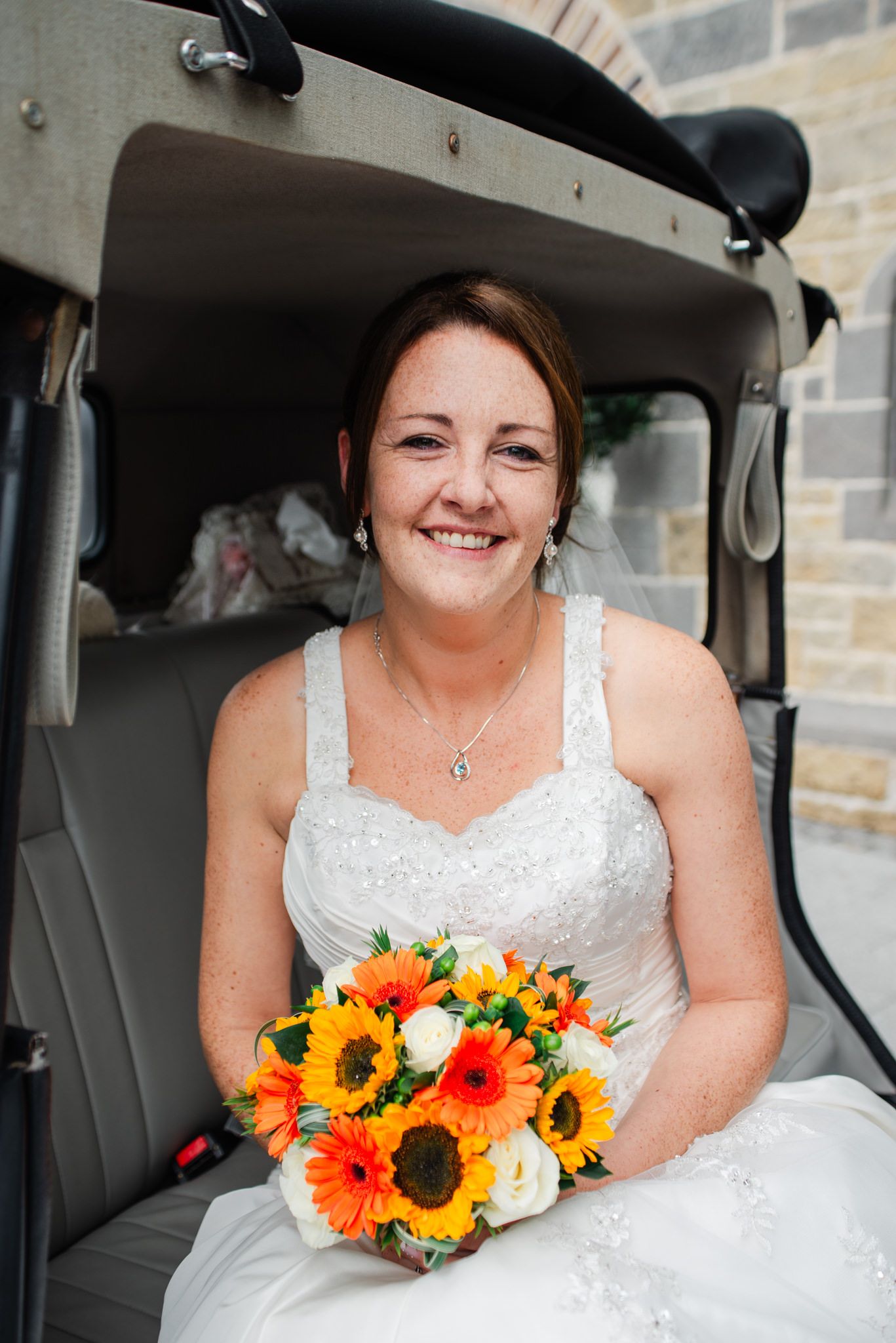 Bride with sunflower and orange floral bouquet poses in wedding car.