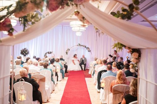 An elegant indoor wedding ceremony venue with white draping, a red carpet aisle, and guests seated in rows under crystal chandeliers.