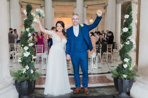 Newlyweds celebrate at ceremonial exit with decorative floral arrangements and white chairs in elegant outdoor venue setting.