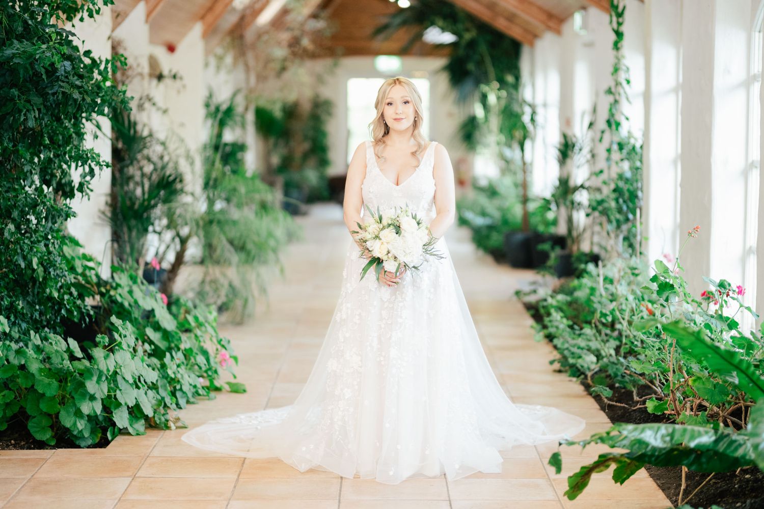 Bride wearing lace wedding gown poses in sunlit greenhouse corridor with white bouquet and green potted plants.