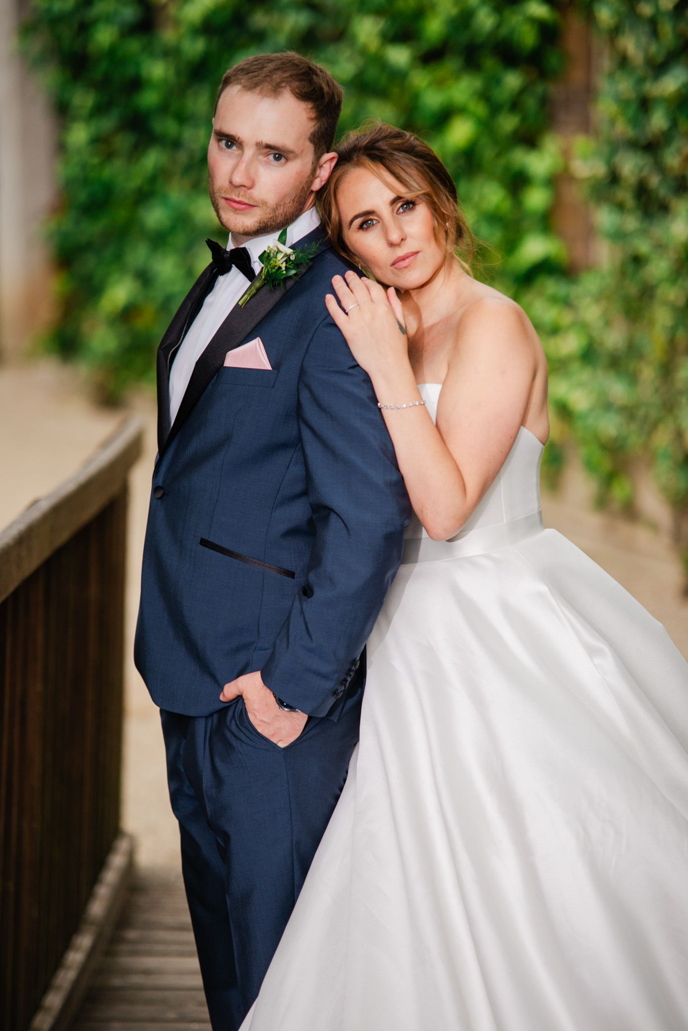 Couple poses together on wooden bridge with greenery backdrop, bride in white ballgown and groom in navy suit.