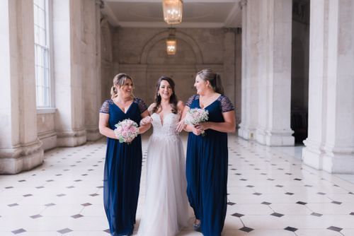 Three figures in formal attire share a candid moment together in an elegant hallway with white marble columns and flooring.