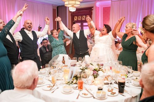 Wedding reception guests celebrate with raised arms at a formal dinner table decorated with floral centerpieces and pink lighting.