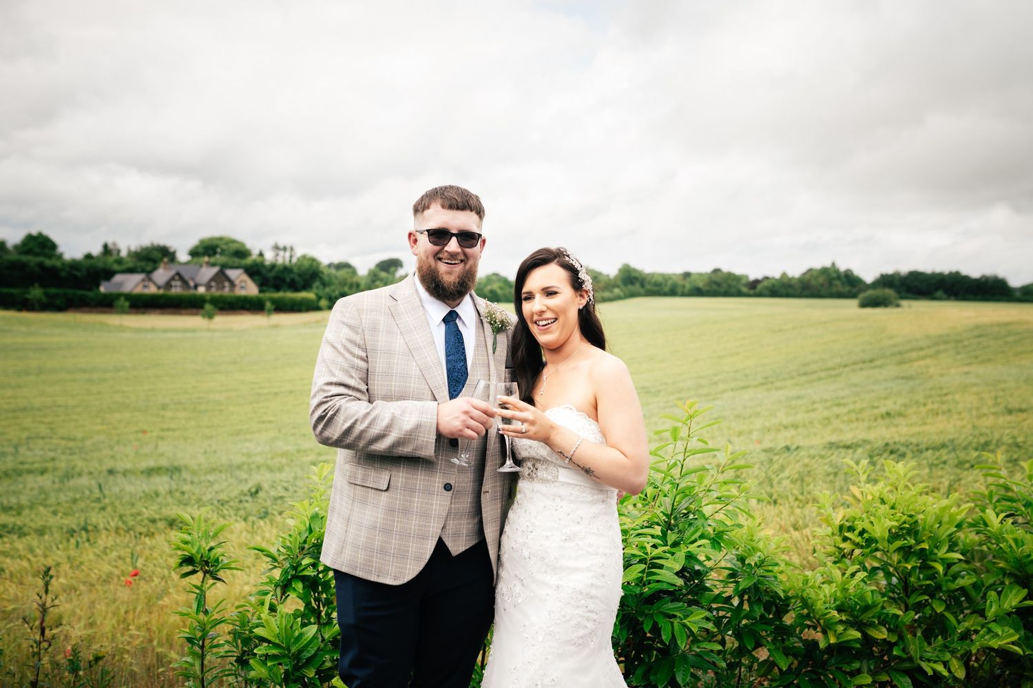 A couple celebrates in a green countryside field while holding champagne glasses on their wedding day.