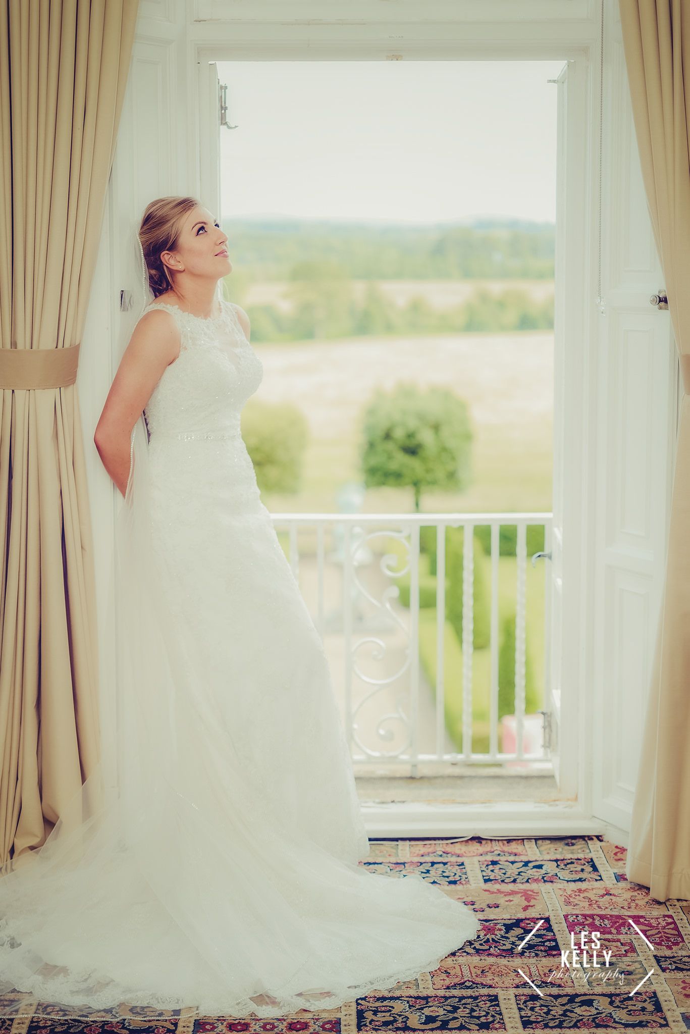 Romantic window portrait of a bride in flowing white gown against countryside view.