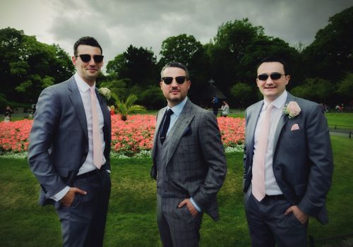 Three men in gray suits and sunglasses pose together in front of a colorful flower garden on an overcast day.