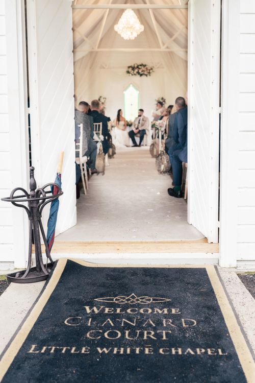 Welcome mat at entrance of Little White Chapel with wedding ceremony taking place in background.