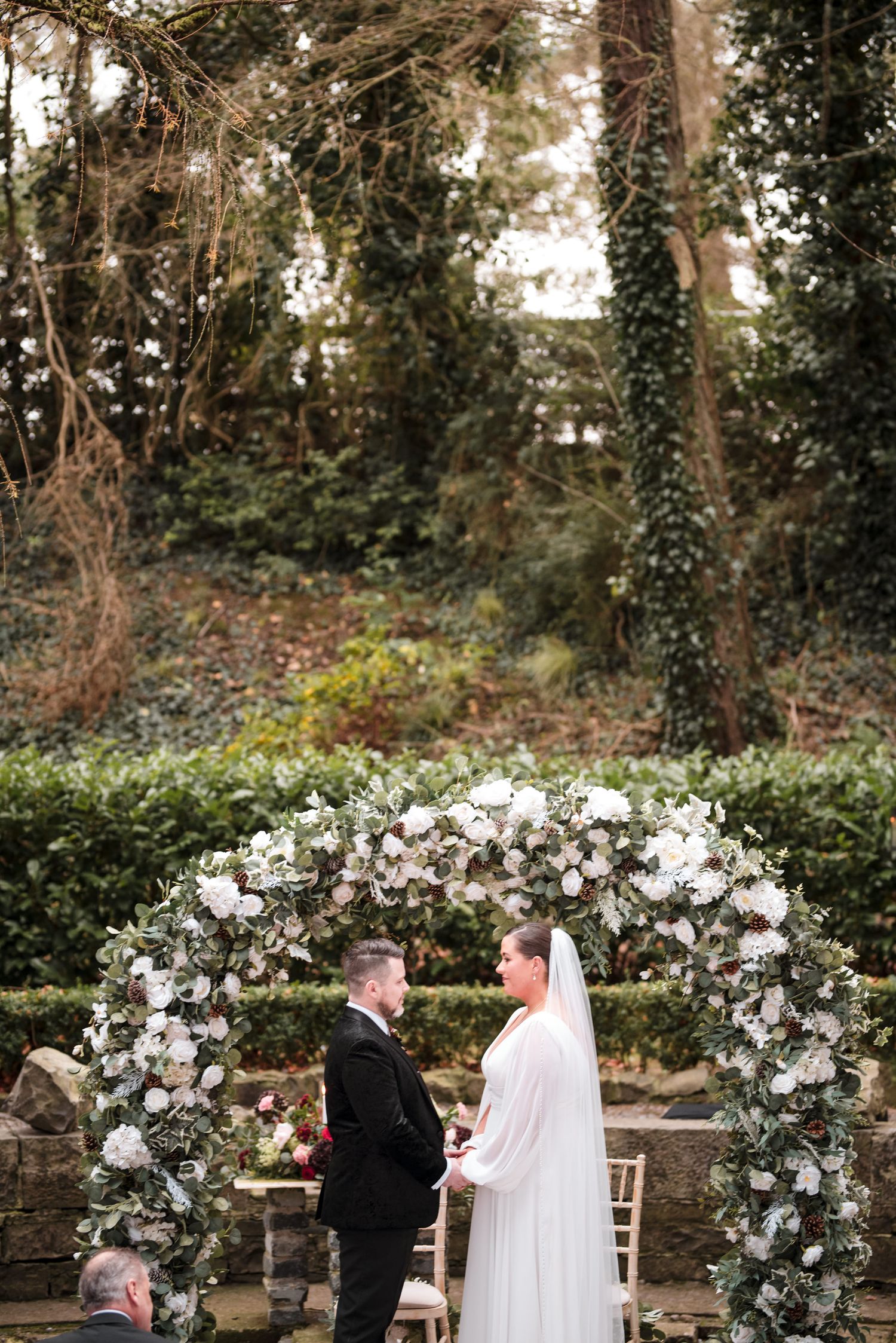 A beautiful wedding ceremony takes place under an elegant white floral arch in a lush garden setting.