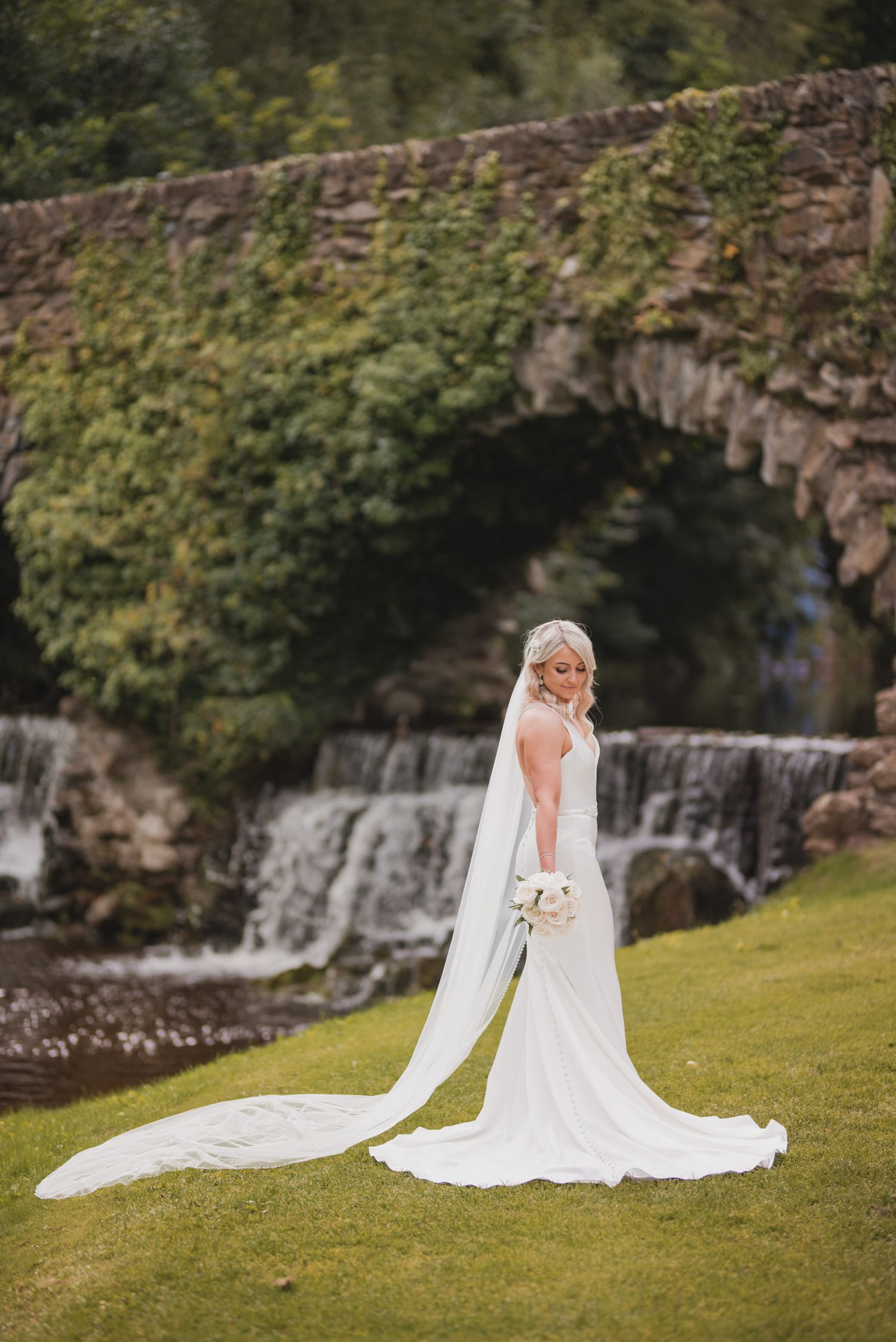 A beautiful bride in a flowing white wedding gown poses gracefully on a grassy hill near a stone bridge.
