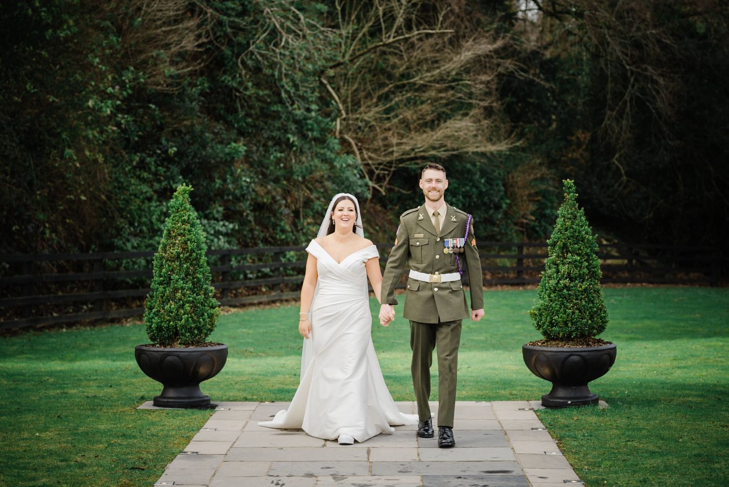 Bride in white dress and groom in military uniform walking together on garden path with topiary.