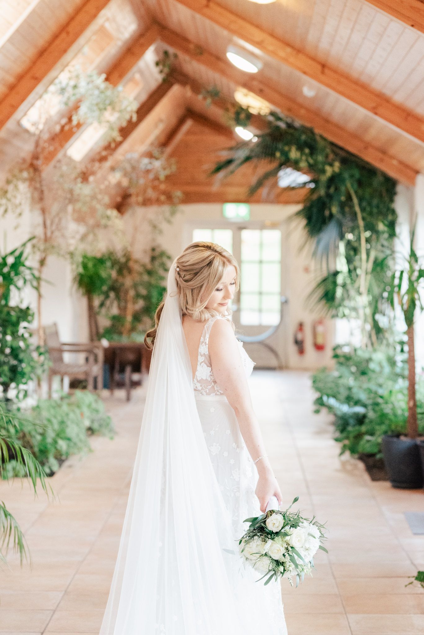Elegant bride in lace dress with long veil holds white bouquet in sunlit greenhouse conservatory filled with greenery.